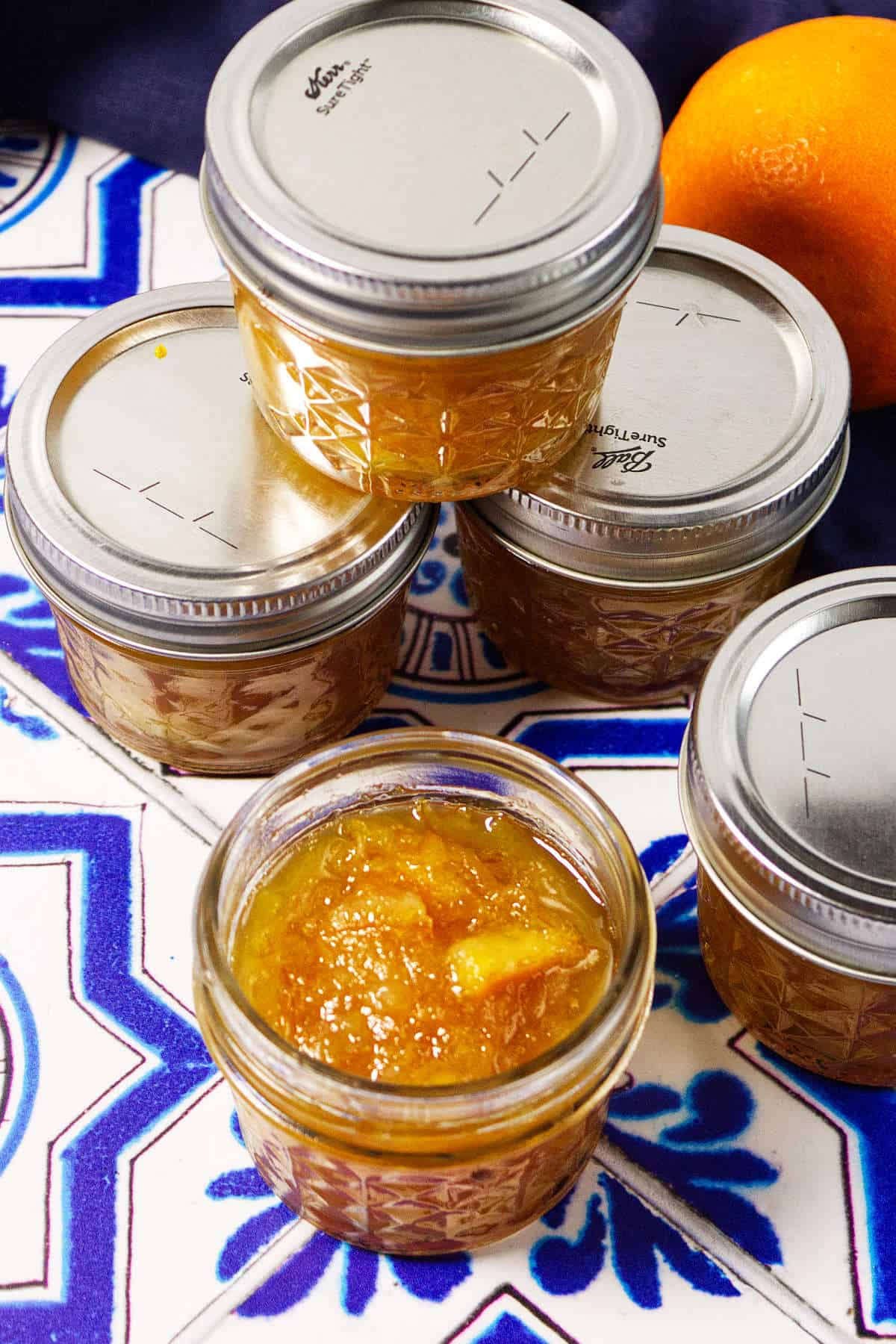 Five small glass jars filled with orange marmalade are arranged on a blue and white patterned tile surface. Four jars are sealed with metal lids, while one is open to reveal the vibrant orange marmalade inside. An orange is partially visible.