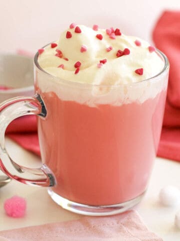 A clear glass mug filled with Pink Velvet hot chocolate, topped with whipped cream and small pink and red heart-shaped sprinkles, sits on a table with a pink napkin and red cloth in the background.