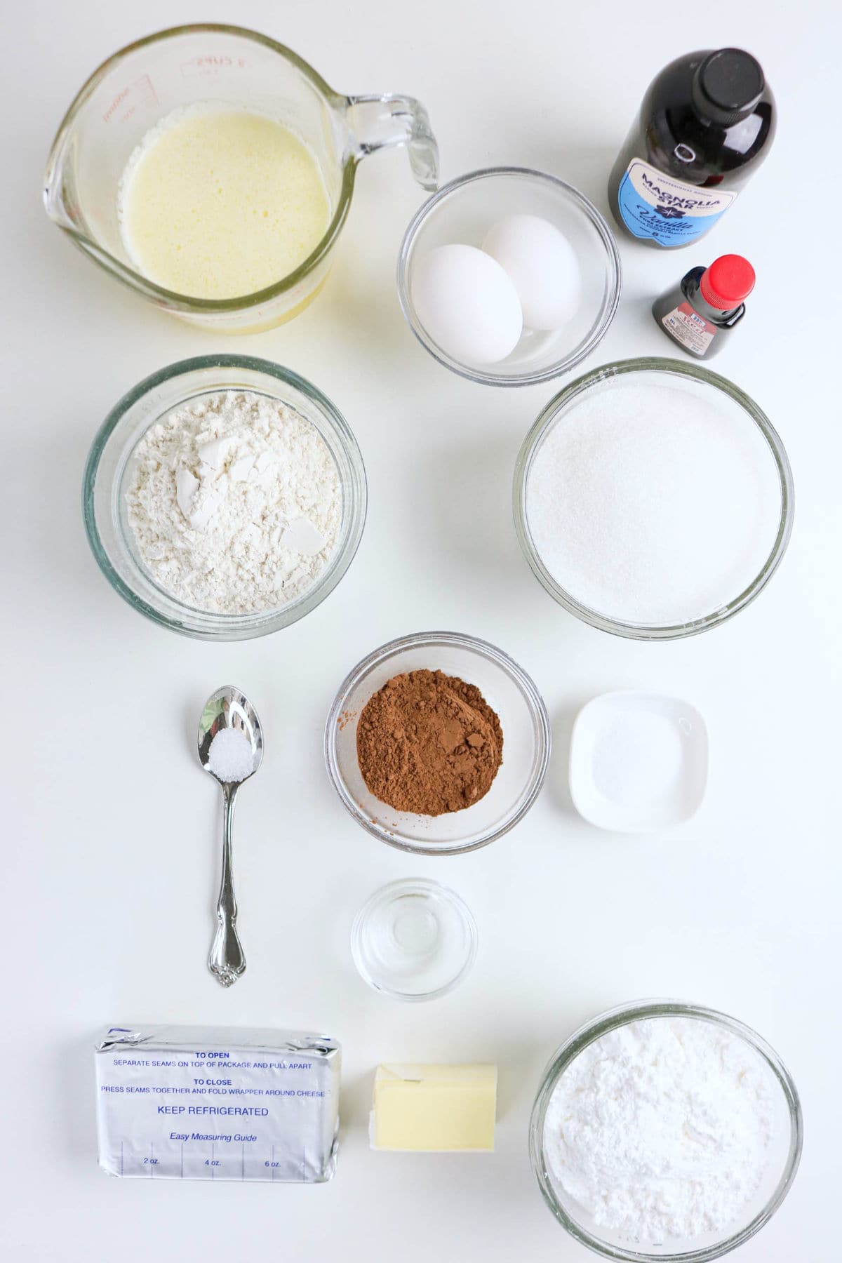 A top-down view of baking ingredients on a white surface, perfect for making Red Velvet Bundt Cake—flour, sugar, cocoa powder, eggs, milk in a measuring cup, butter, baking soda on a spoon, vanilla, red food coloring, and powdered sugar.