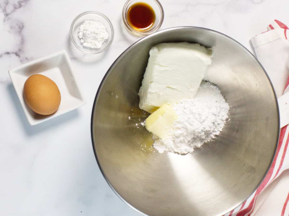 A metal mixing bowl with cream cheese, butter, and powdered sugar sits ready for a Red Velvet Bundt Cake. Nearby are a brown egg in a white dish, plus small bowls of cornstarch and vanilla extract on a white marble surface.