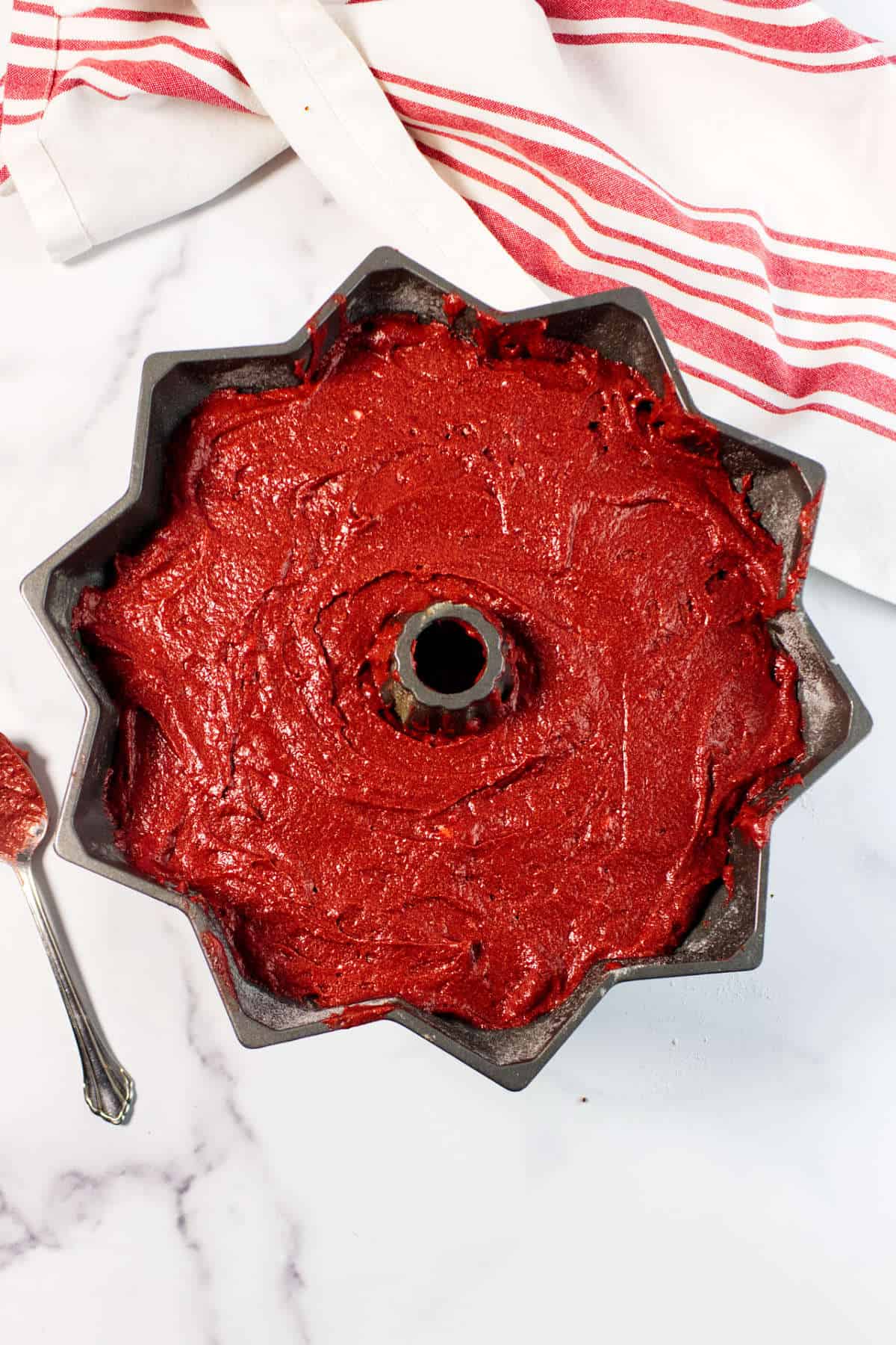 A bundt pan filled with unbaked Red Velvet Bundt Cake batter sits on a white marble surface, next to a spoon and a white kitchen towel with red stripes.