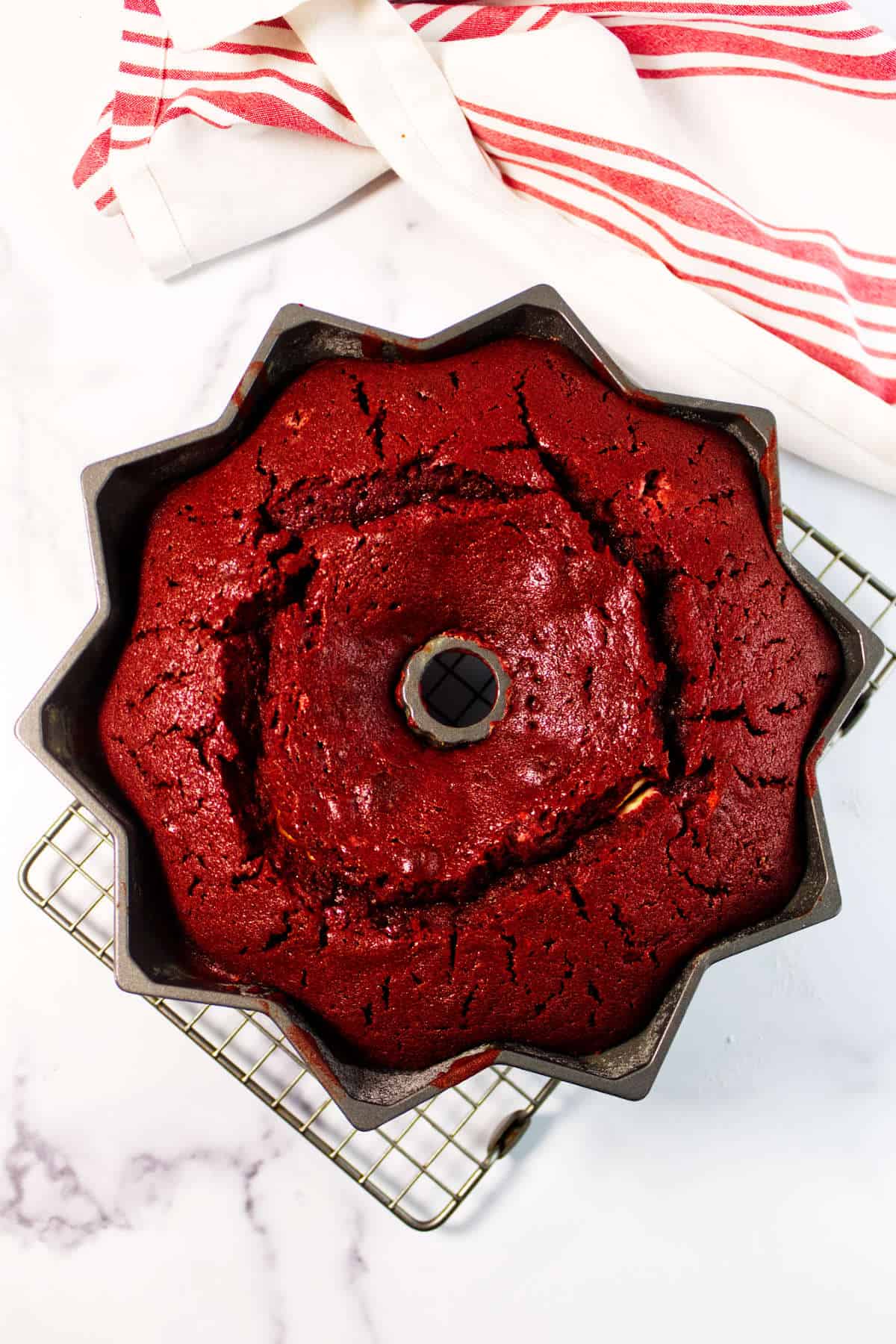 A freshly baked Red Velvet Bundt Cake sits in a pan on a cooling rack, with a white and red striped kitchen towel nearby on the marble surface.