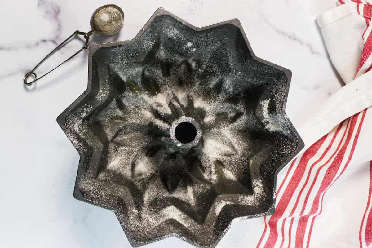 A fluted Bundt cake pan dusted with flour sits on a white surface, ready to bake a classic Red Velvet Bundt Cake, next to a metal ice cream scoop and a red-and-white striped dish towel.