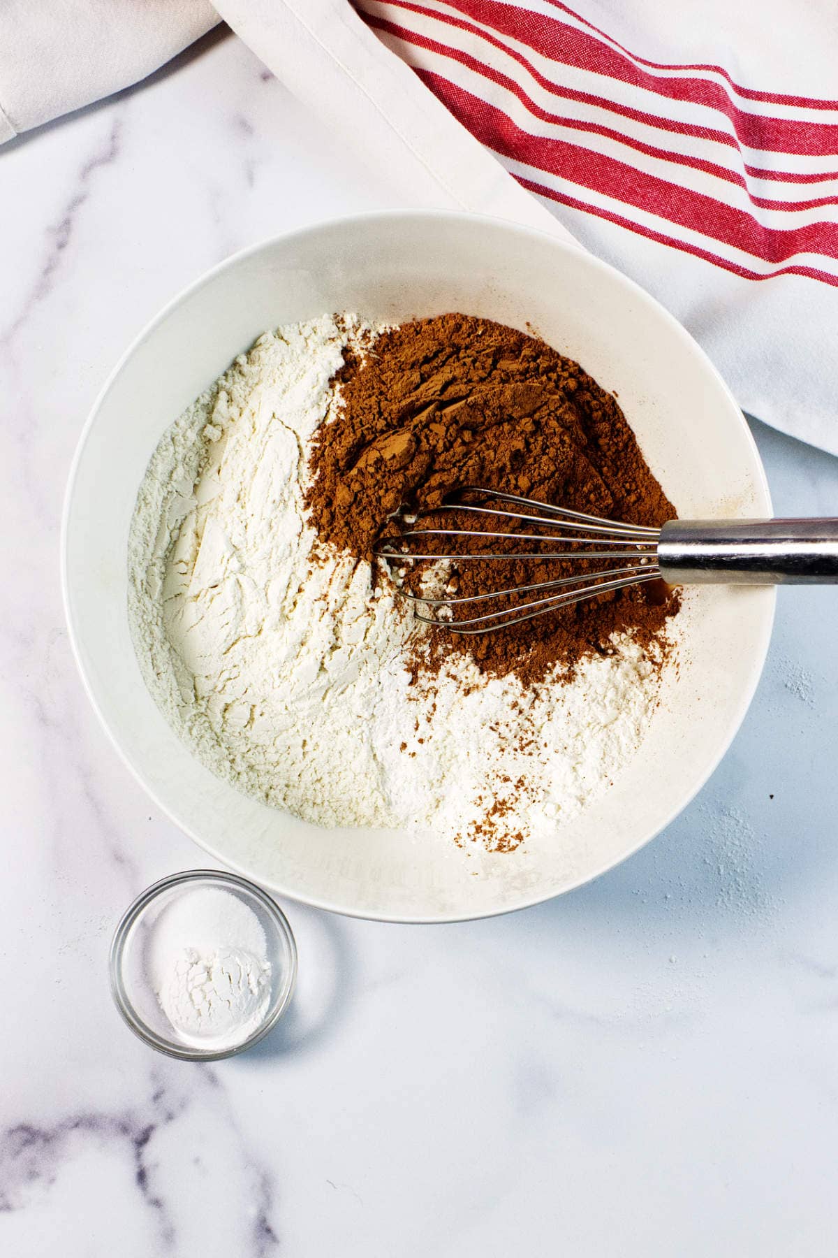A white bowl with flour and cocoa powder being whisked together, perfect for making Red Velvet Bundt Cake, sits on a marble surface next to a small bowl of baking powder and a white towel with red stripes.
