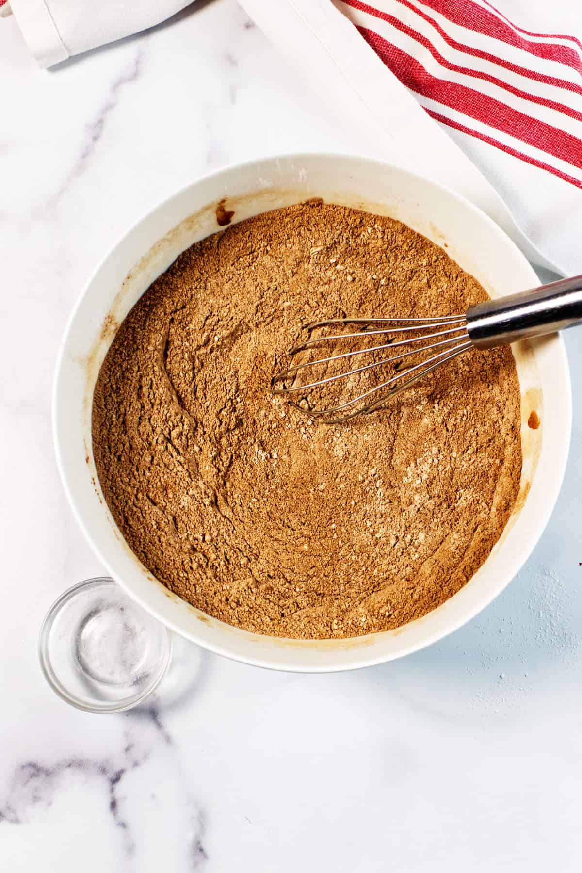 A white mixing bowl filled with brown dry ingredients for a Red Velvet Bundt Cake is being whisked, sitting on a white countertop next to a red-and-white striped kitchen towel and a small empty glass bowl.