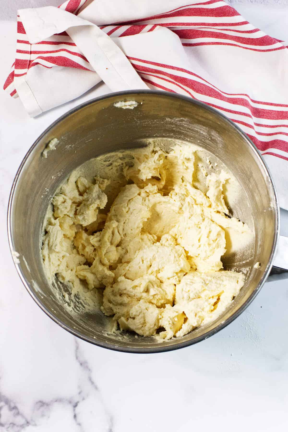 A stainless steel mixing bowl filled with creamy, pale cookie dough sits on a white marble surface, ready to be transformed into a delicious Red Velvet Bundt Cake, next to a white kitchen towel with red stripes.