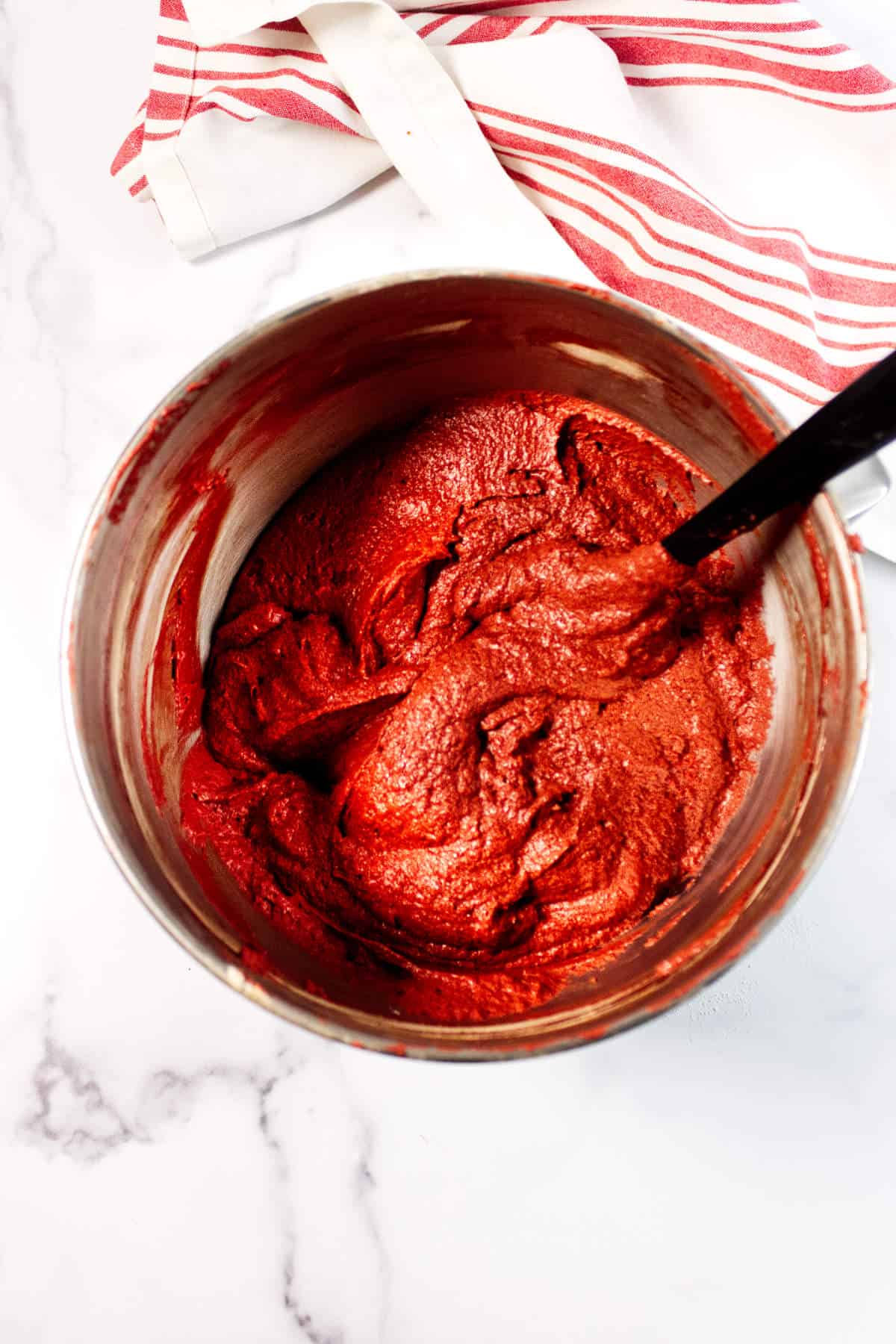 A metal mixing bowl filled with red cake batter for Red Velvet Bundt Cake is being stirred with a black spatula, set on a white marble surface with a red and white striped kitchen towel nearby.