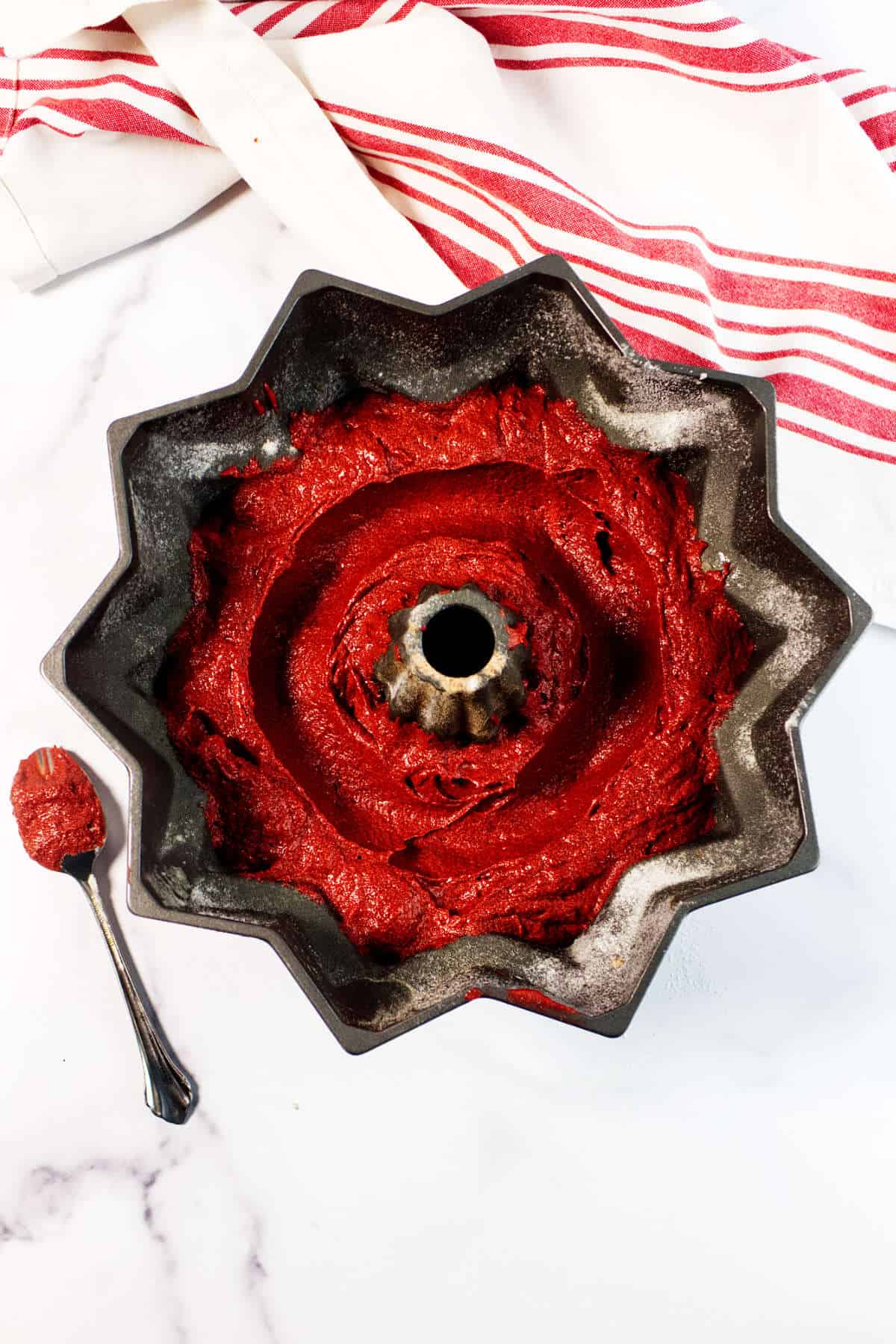 A bundt pan filled with Red Velvet Bundt Cake batter sits on a white surface next to a spoon with batter on it. A white towel with red stripes is partially visible in the background.