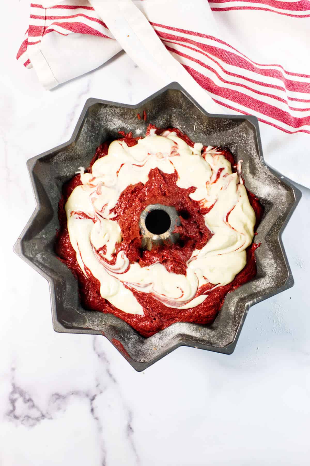A Red Velvet Bundt Cake pan filled with swirled red and white batter sits on a marble surface next to a white and red striped kitchen towel.