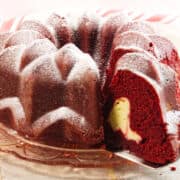 A Red Velvet Bundt Cake dusted with powdered sugar sits on a glass plate. One slice has been cut, revealing a creamy cheesecake filling inside this decadent dessert.