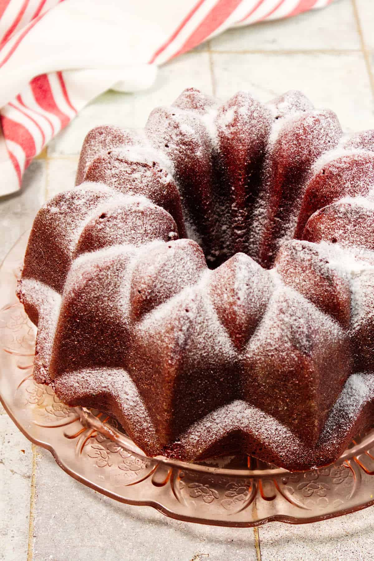 A Red Velvet Bundt Cake dusted with powdered sugar sits on a vintage glass plate, with a red and white striped kitchen towel in the background.