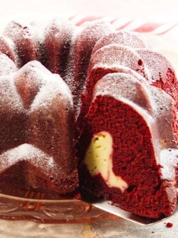 A Red Velvet Bundt Cake dusted with powdered sugar, with a slice removed to reveal a creamy cheesecake filling inside, displayed elegantly on a glass plate.