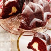 A Red Velvet Bundt Cake with a creamy cheesecake filling is displayed on a glass cake stand. One slice of the decadent Red Velvet Bundt Cake is cut and placed on a white plate with a gold rim in the foreground.