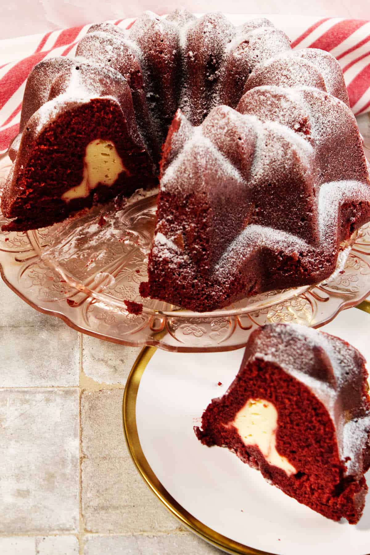 A Red Velvet Bundt Cake with a creamy cheesecake filling is displayed on a glass cake stand. One slice of the decadent Red Velvet Bundt Cake is cut and placed on a white plate with a gold rim in the foreground.