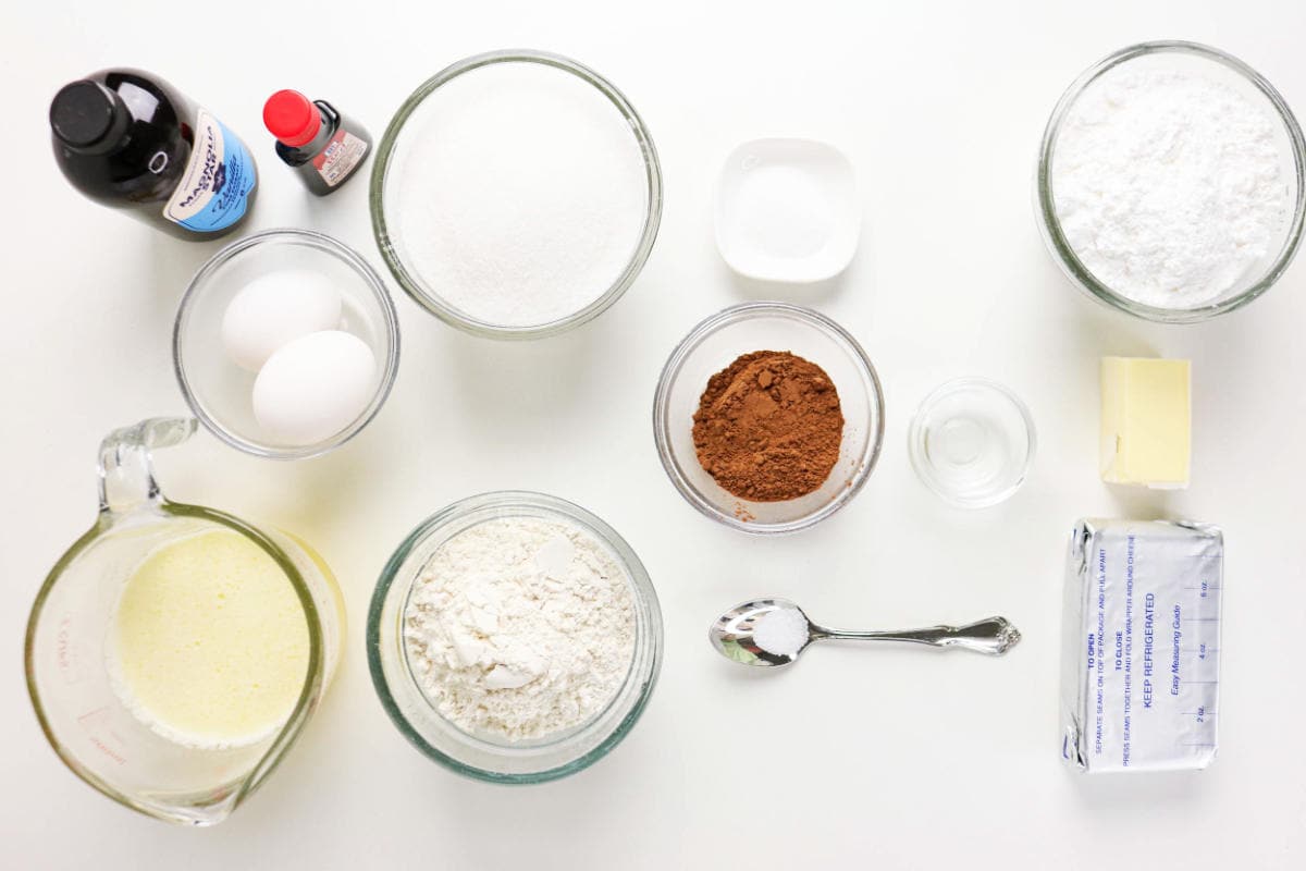Various baking ingredients are arranged on a white surface, ready for red velvet cake brownies, including eggs, sugar, flour, cocoa powder, baking soda, butter, cream cheese, vanilla extract, and a measuring cup with a liquid mixture.