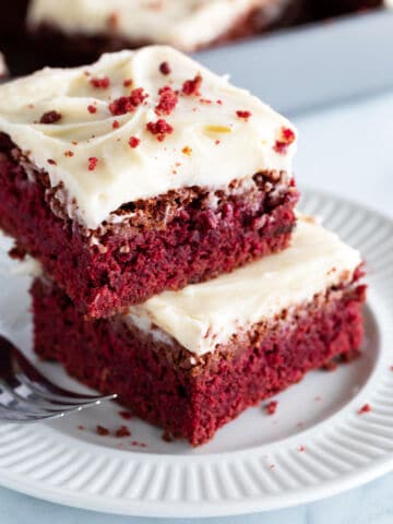 Two pieces of red velvet cake brownies with cream cheese frosting are stacked on a white plate, with a fork beside them. The brownies are moist and red, topped with smooth white frosting and sprinkled with red crumbs.