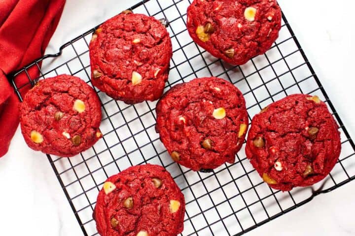 Six red velvet chocolate chip cookies with white and dark chocolate chips cool on a black wire rack, with a red cloth partially visible in the corner on a white surface.