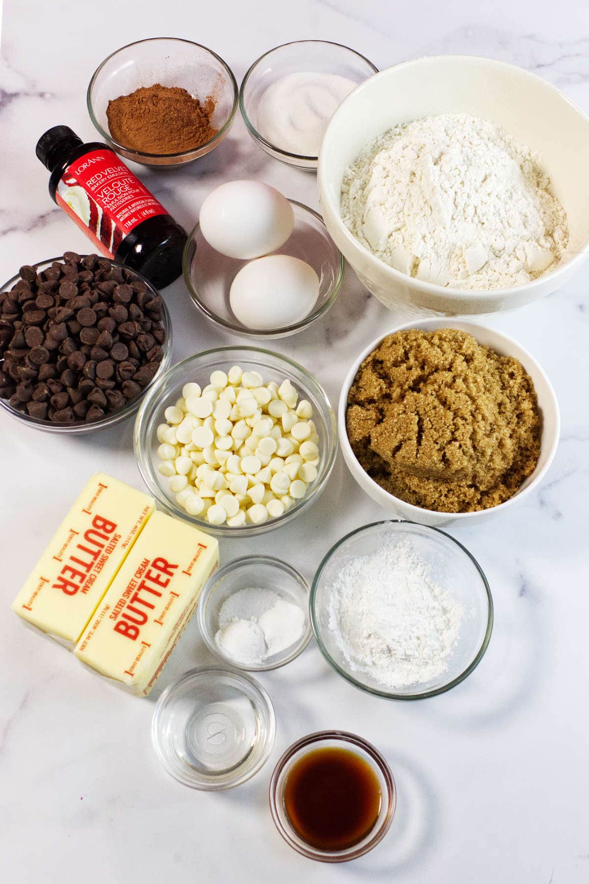 Assorted baking ingredients in bowls on a white surface, perfect for making red velvet chocolate chip cookies—includes flour, brown sugar, sugars, cocoa powder, chocolate chips, white chocolate chips, eggs, butter sticks, baking powder, vanilla extract, and soda.