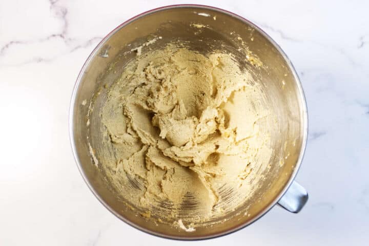 A metal mixing bowl containing creamy, light brown red velvet chocolate chip cookie dough sits on a white marble countertop. The dough appears freshly mixed and smooth.
