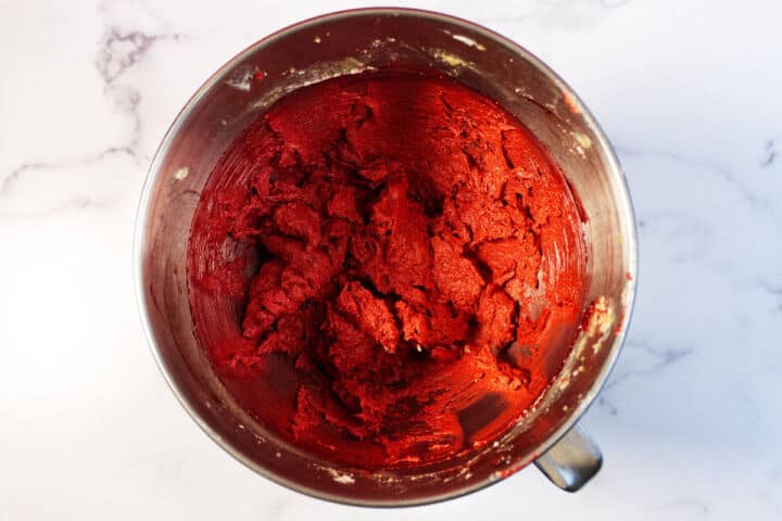 A metal mixing bowl filled with bright red velvet chocolate chip cookie dough sits on a white marble surface, with some dough smeared on the sides of the bowl.