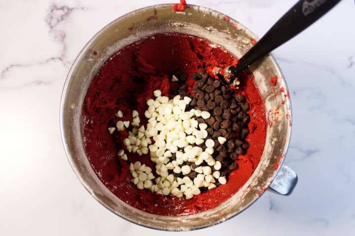 A mixing bowl filled with red velvet chocolate chip cookie dough, topped with white and dark chocolate chips, and a black spatula resting in the bowl, on a white marble surface.