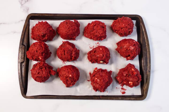 A baking tray lined with parchment paper holds twelve scoops of red velvet chocolate chip cookie dough, spaced apart and ready to be baked. The background is a white marble surface.