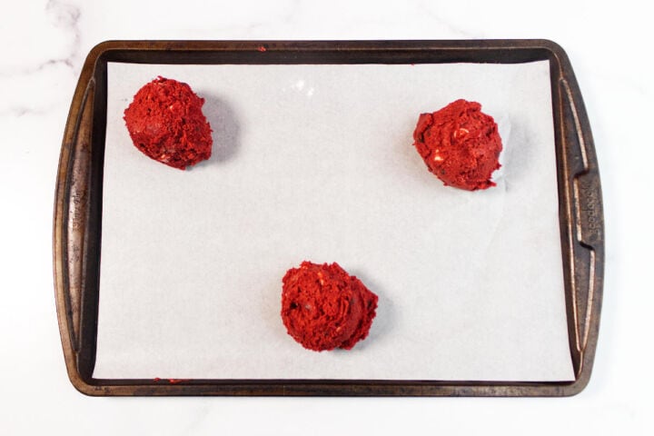 Three scoops of red velvet chocolate chip cookie dough are spaced out on a parchment-lined baking sheet, ready to be baked. The baking sheet sits on a white surface.