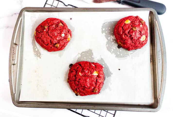 Three large red velvet chocolate chip cookies with white and dark chips rest on a parchment-lined baking sheet. The cookies are spaced apart, and the sheet sits atop a black cooling rack.