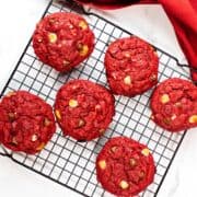 Six red velvet chocolate chip cookies with white and milk chocolate chips are cooling on a black wire rack. A red cloth napkin peeks from the corner, set against a white marble surface.