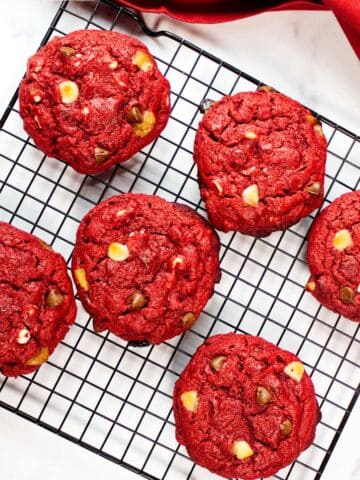Six red velvet chocolate chip cookies with white and milk chocolate chips are cooling on a black wire rack. A red cloth napkin peeks from the corner, set against a white marble surface.