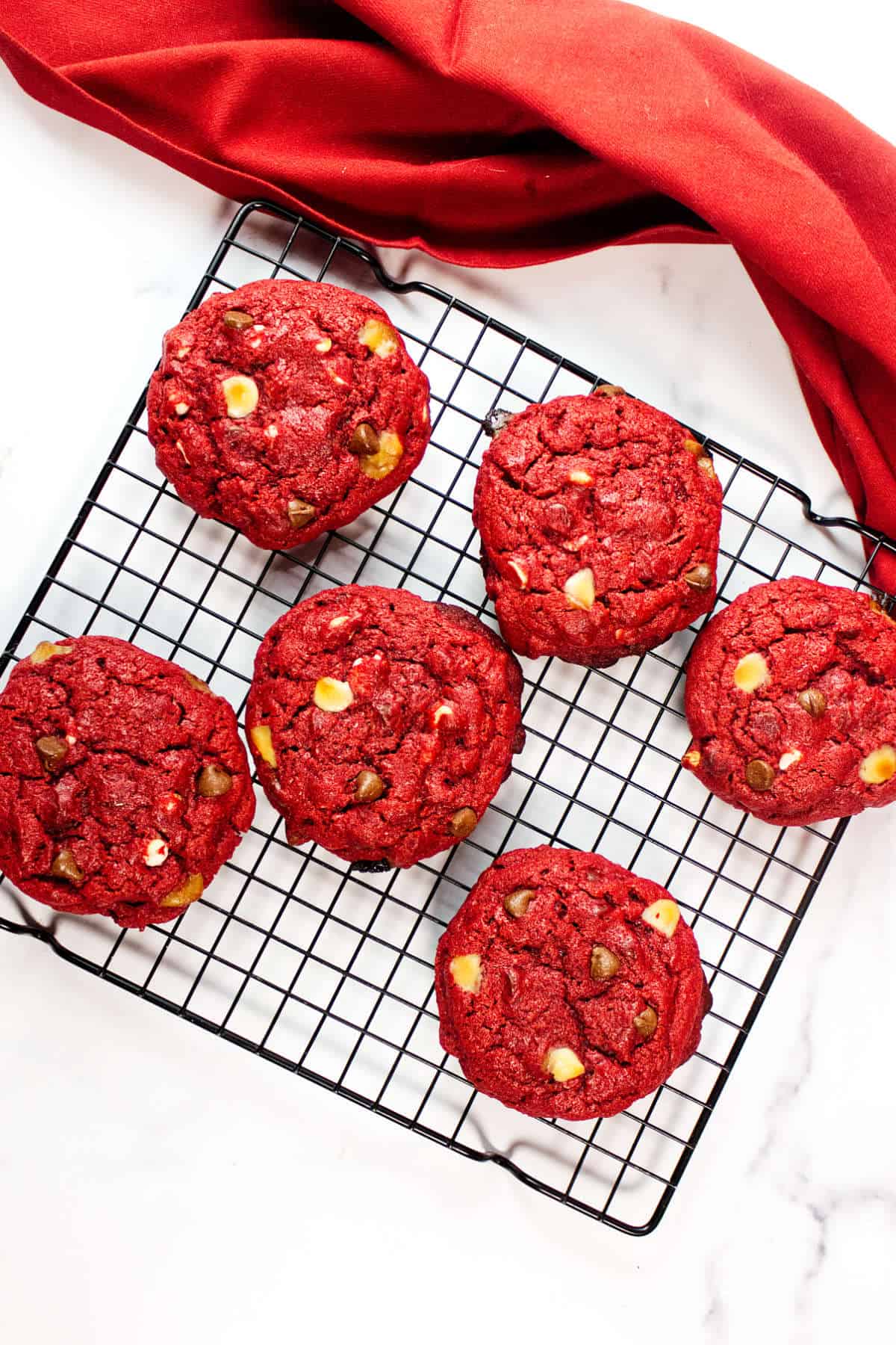 Six red velvet chocolate chip cookies with white and dark chocolate chips cooling on a black wire rack, with a red cloth partially visible in the background on a white surface.