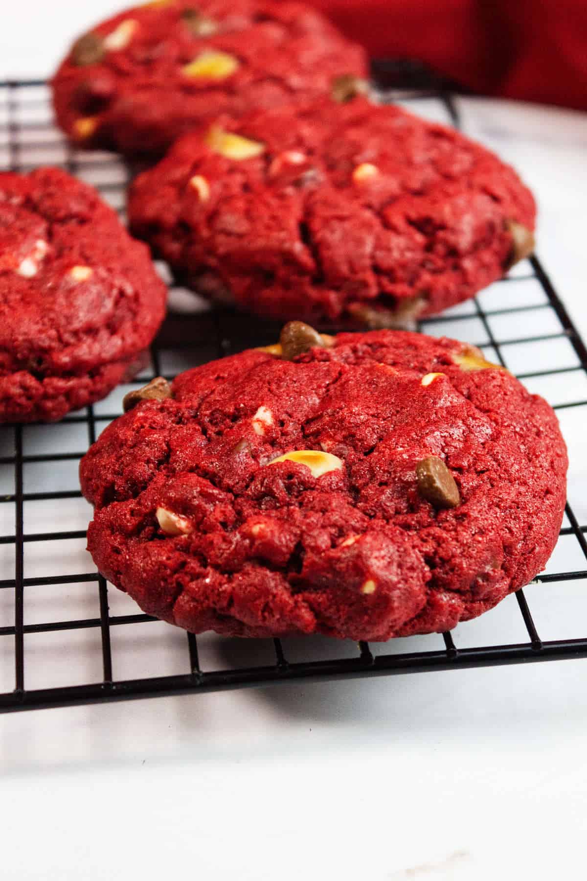 Close-up of freshly baked red velvet chocolate chip cookies, their thick texture packed with white and chocolate chips, cooling on a black wire rack against a white background.