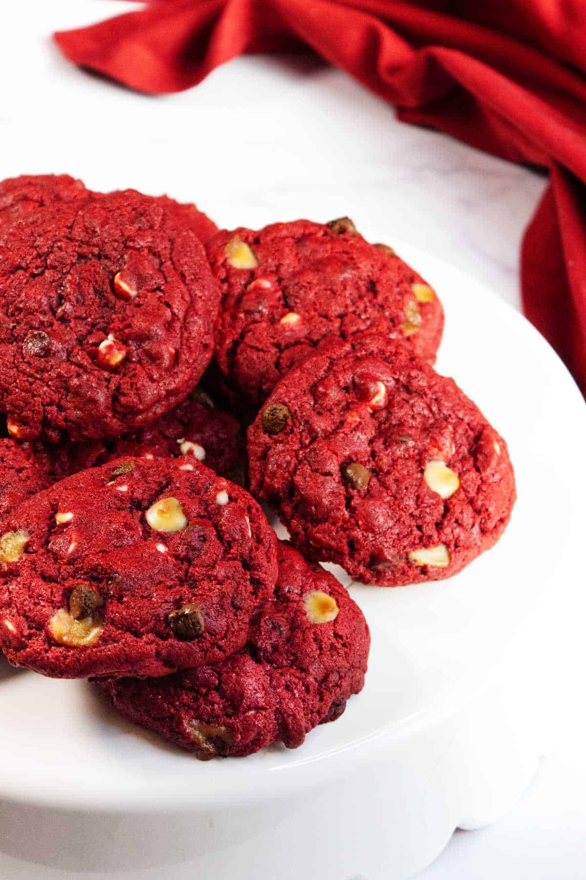 A plate of vibrant red velvet chocolate chip cookies with white and dark chips sits on a white cake stand, with a red cloth draped in the background.
