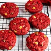 Six vibrant red velvet chocolate chip cookies, studded with white and milk chocolate chips, rest on a black cooling rack beside a red cloth on a white surface.