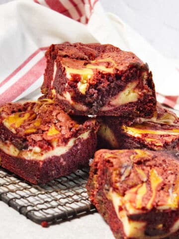 Four Red Velvet Cream Cheese Brownies with creamy swirls are stacked on a black cooling rack, with a white and red striped cloth in the background. The brownies look rich and moist, showcasing their distinct marbled patterns.