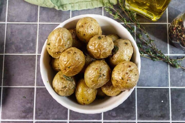 A white bowl filled with roasted baby potatoes, garnished with herbs, sits on a tiled surface next to a sprig of thyme, a green cloth, and a bottle of olive oil.