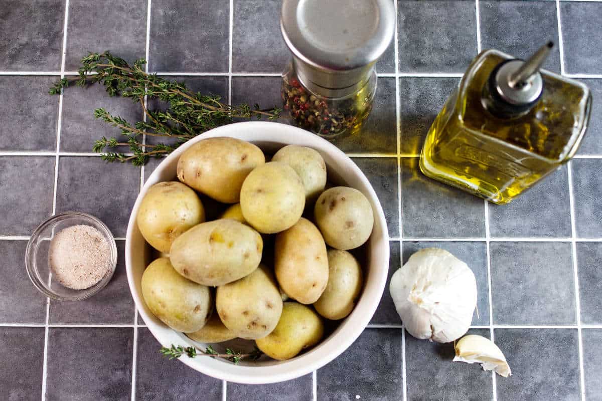A bowl of small potatoes, a sprig of thyme, a garlic bulb, a small dish of salt, a pepper grinder, and a bottle of olive oil arranged on a gray tiled countertop.