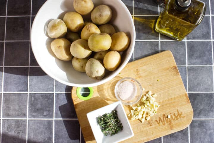 A white bowl of small potatoes, a glass bottle of olive oil, and a wooden cutting board with chopped garlic, salt in a small bowl, and fresh herbs sit on a tiled kitchen countertop in sunlight.