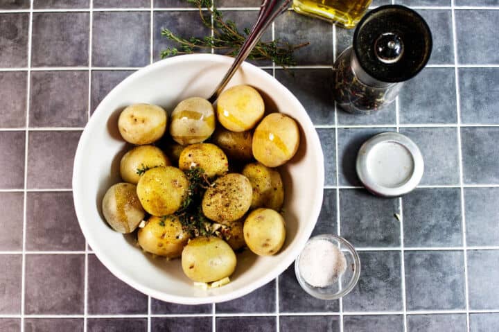 A white bowl filled with small potatoes, topped with herbs and spices, sits on a gray tiled surface. Nearby are a pepper grinder, an open salt container, a small glass of salt, and a bottle of oil.
