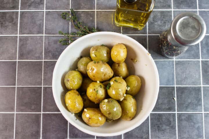 A white bowl filled with seasoned baby potatoes sits on a gray tiled surface, with fresh herbs, a bottle of olive oil, and a pepper grinder nearby.