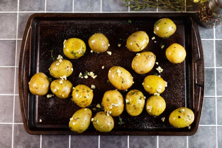 Small whole potatoes coated with chopped herbs and garlic are arranged on a dark baking tray, ready for roasting. The tray sits on a gray tiled countertop with sprigs of herbs and seasoning visible nearby.