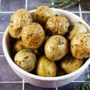 A white bowl filled with roasted baby potatoes, garnished with herbs, sits on a tiled surface. The potatoes have crispy, golden skin with visible seasoning.