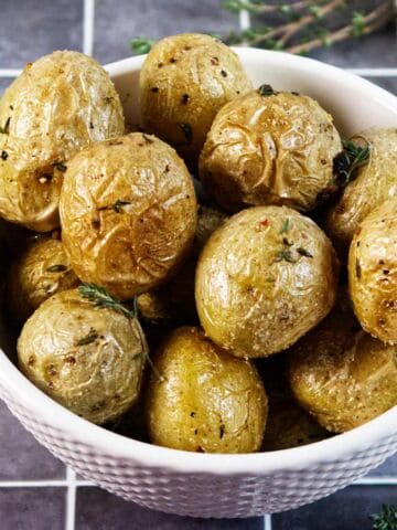 A white bowl filled with roasted baby potatoes, garnished with herbs, sits on a tiled surface. The potatoes have crispy, golden skin with visible seasoning.