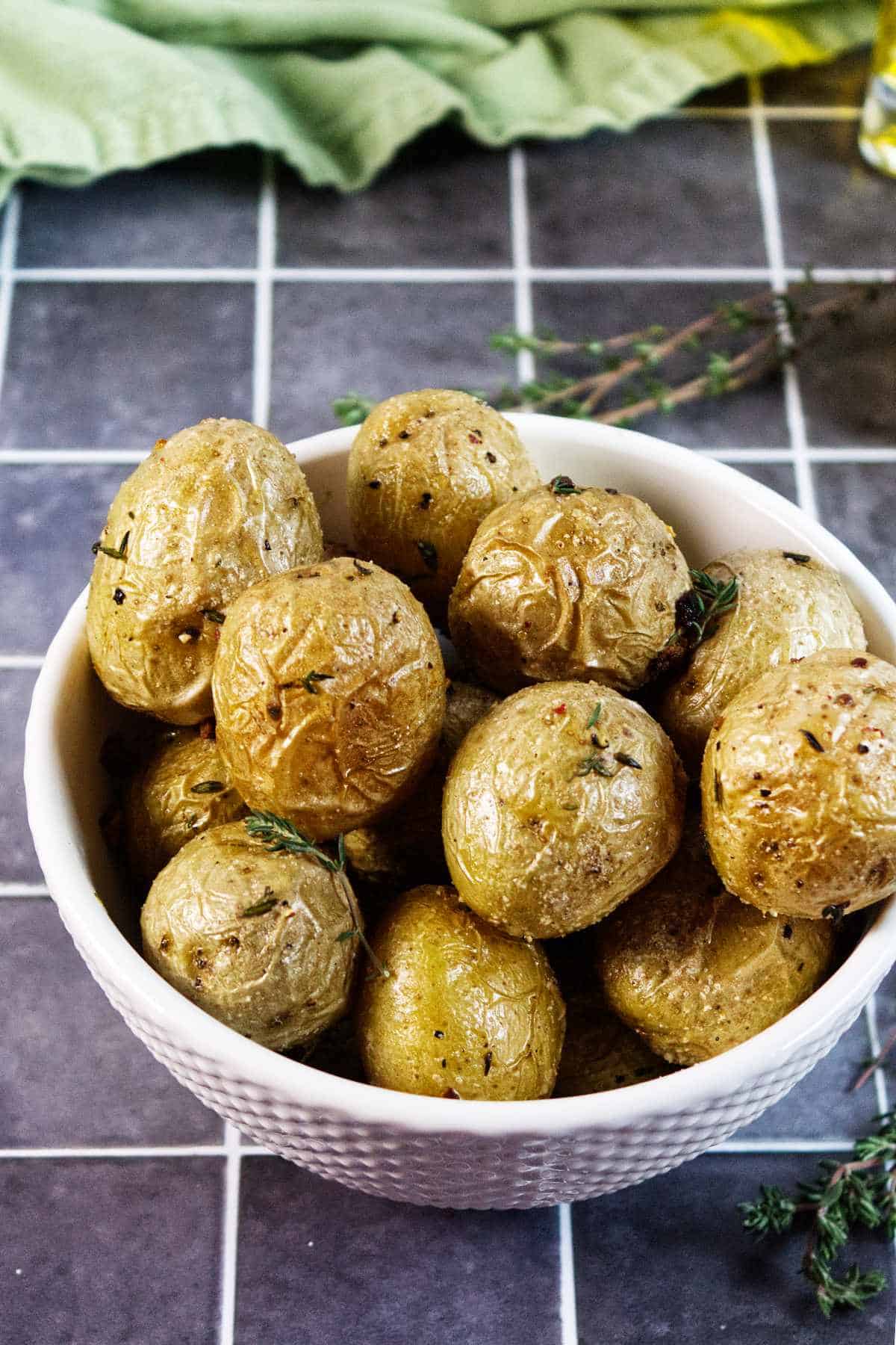 A white bowl filled with roasted baby potatoes, garnished with herbs, sits on a dark tiled surface. A green cloth and sprigs of fresh thyme are visible in the background.