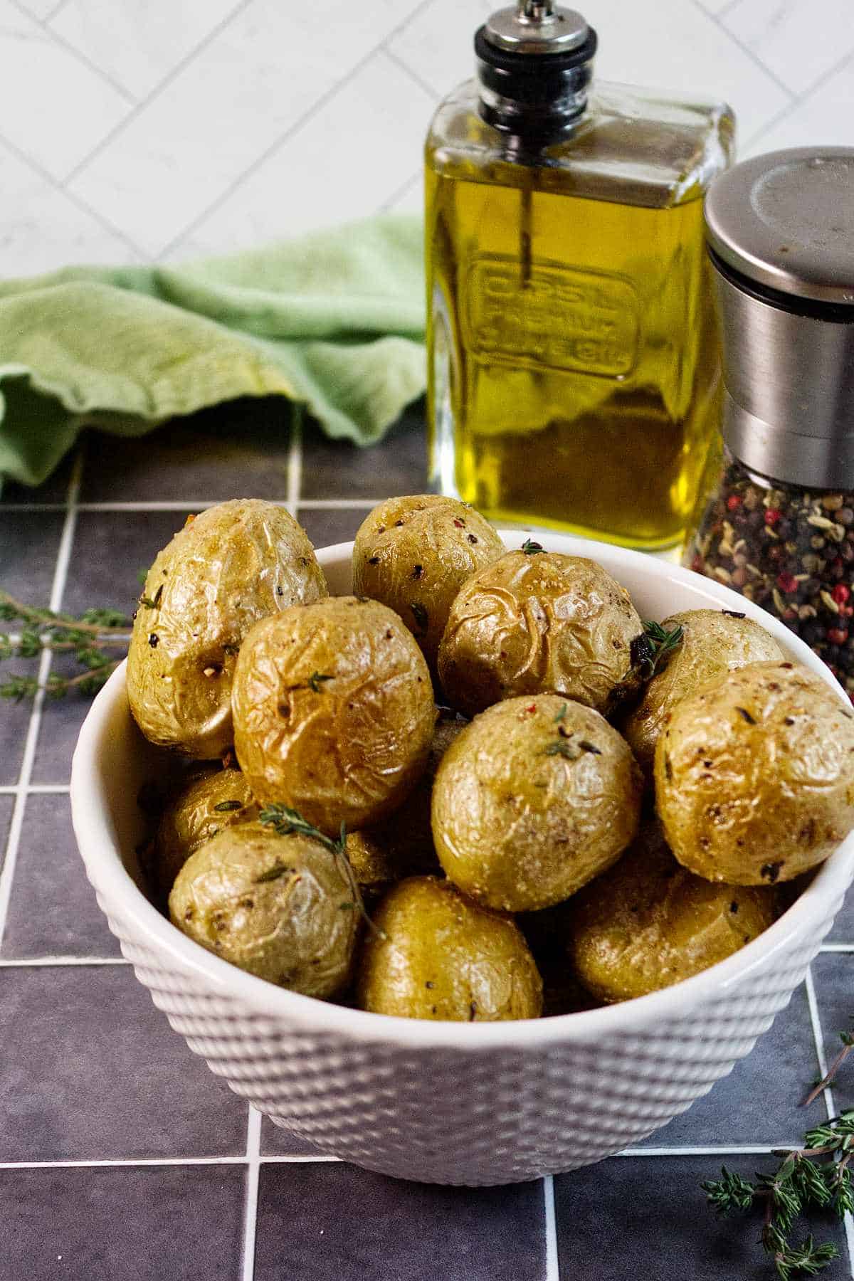 A white bowl filled with roasted baby potatoes garnished with herbs sits on a tiled counter. Behind the bowl are a bottle of olive oil, a pepper grinder, and a green cloth.