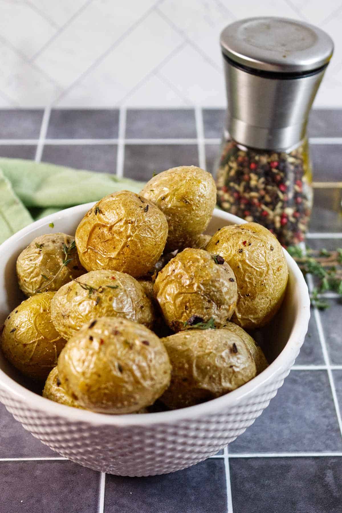 A white bowl filled with roasted baby potatoes seasoned with herbs, placed on a tiled surface with a pepper grinder and a green napkin in the background.