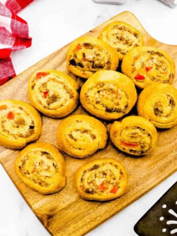 Ten golden-brown sausage crescent roll pinwheels filled with meat and diced red peppers are arranged on a wooden board. A red and white checkered cloth is nearby, with part of a black spatula visible in the corner.