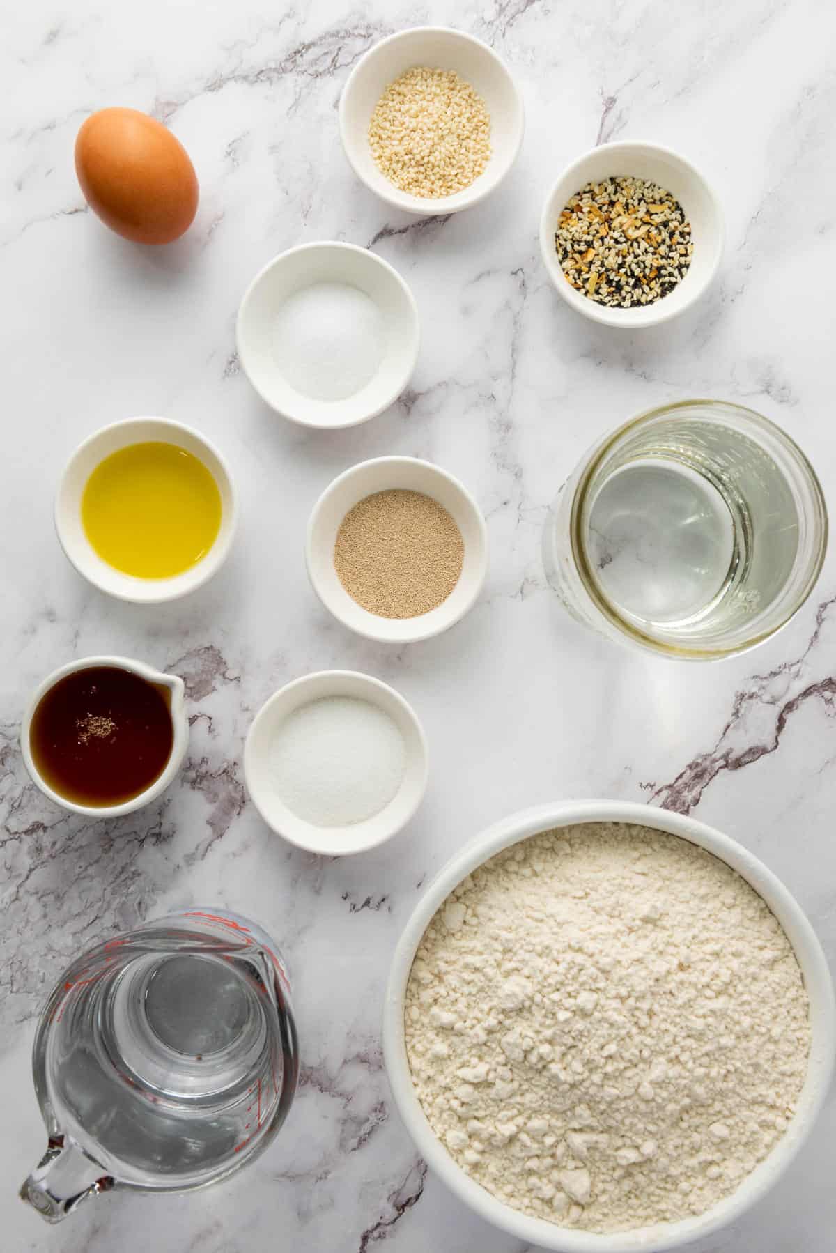 Top-down view of baking ingredients on a marble surface: an egg, bowls of flour, yeast, sugar, salt, olive oil, water, maple syrup, and two small bowls of assorted seeds.