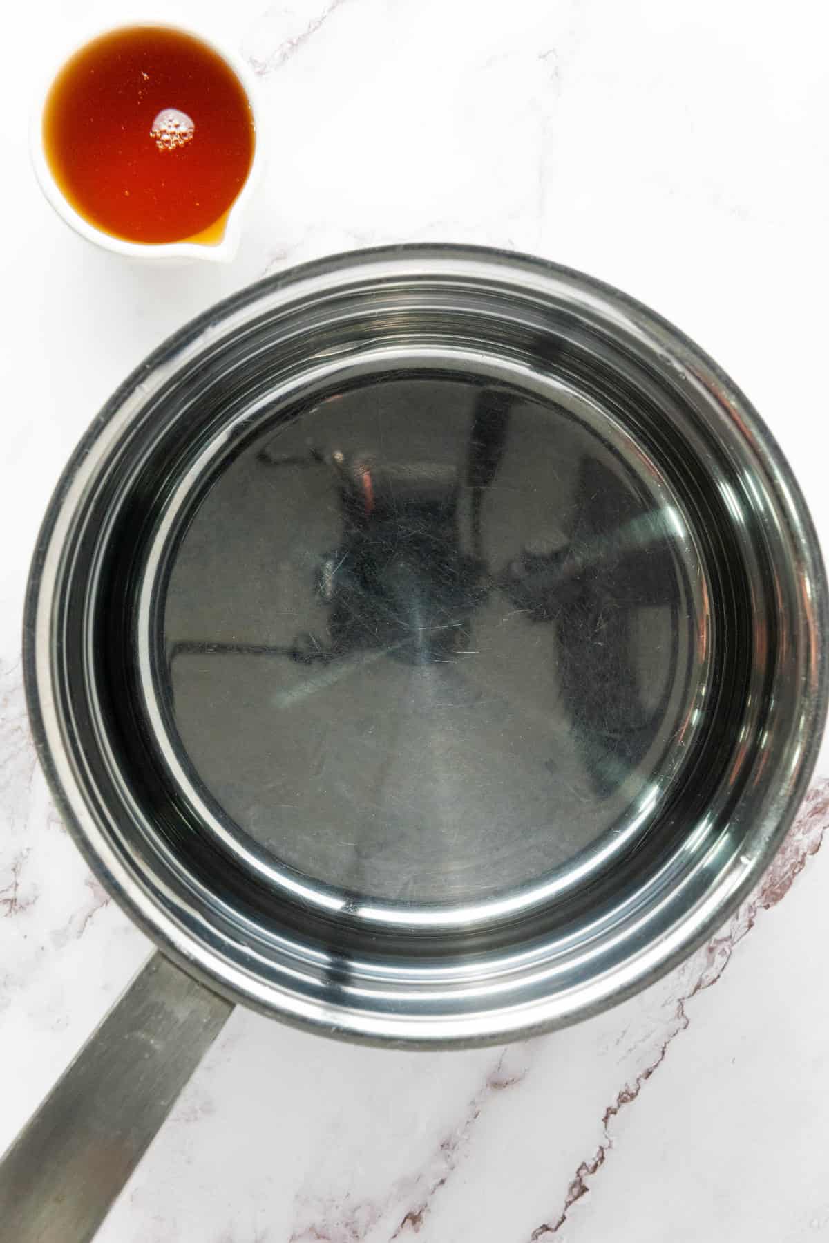 A stainless steel saucepan filled with water sits on a white marble countertop. Beside it is a small white container holding a reddish-brown sauce.