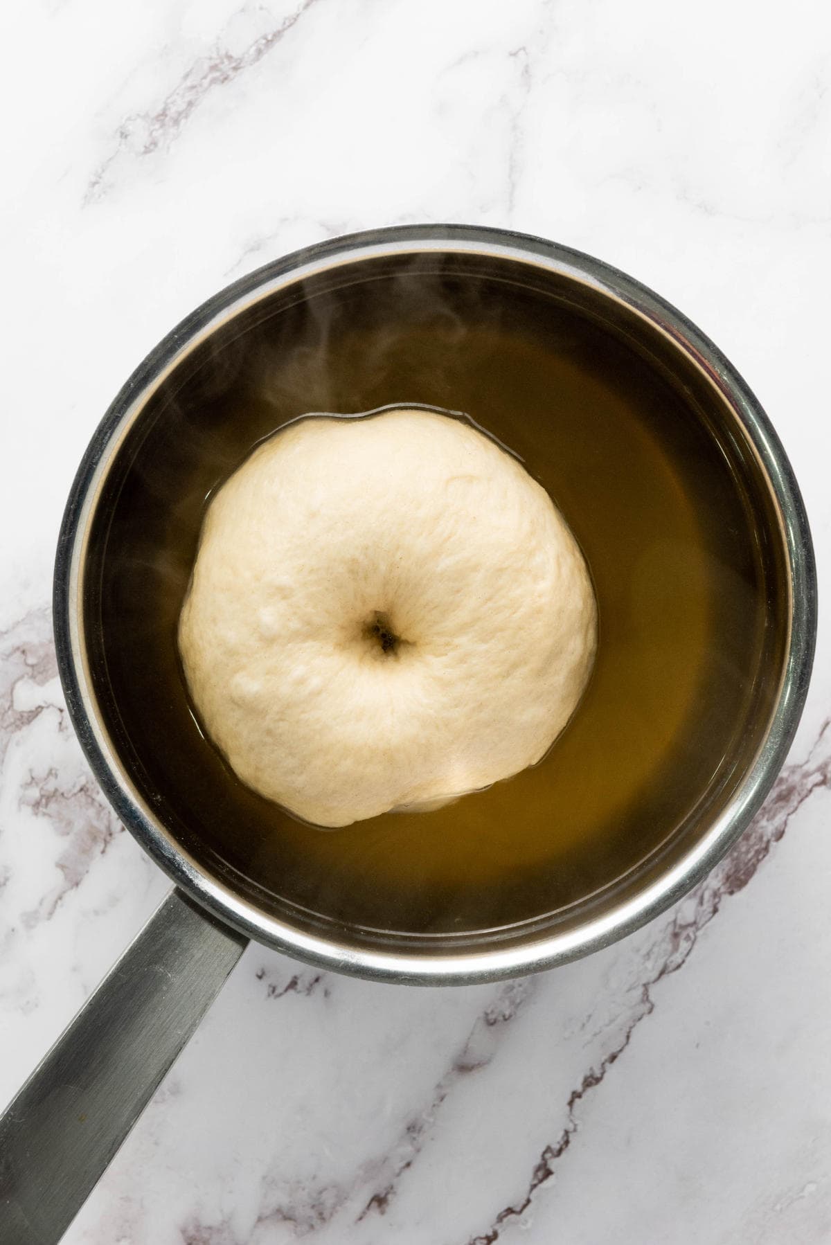 A round piece of dough with a hole in the center is boiling in a pot of water, shown from above on a white marble surface. Steam is rising from the pot.